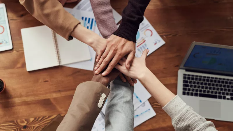 Six hands placed on top of each other over a desk.